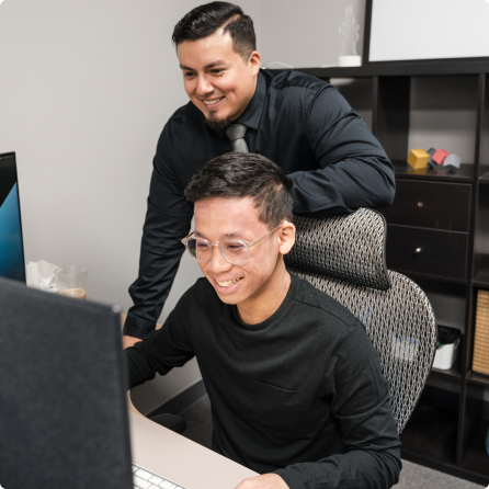 Alejandro standing behind Michael as they both look at computer screens smiling